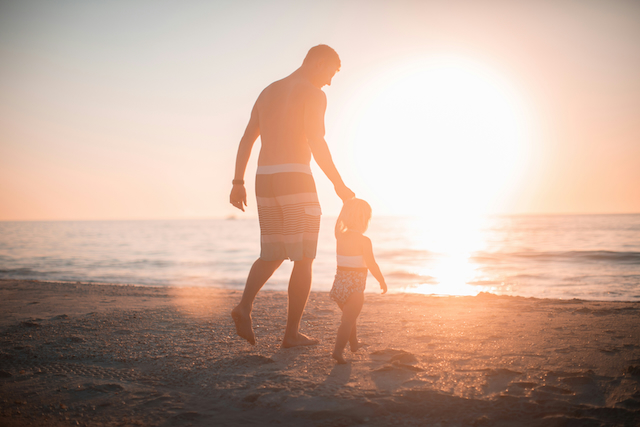 Silhuet af en far og et lille barn, der går hånd i hånd på stranden i solnedgang